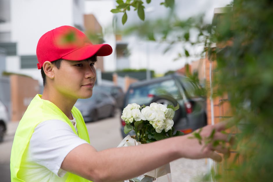 A delivery person wearing a red cap and a bright yellow high-visibility vest is placing a bouquet of fresh white roses and peonies wrapped in transparent cellophane and white paper onto a doorstep, wi