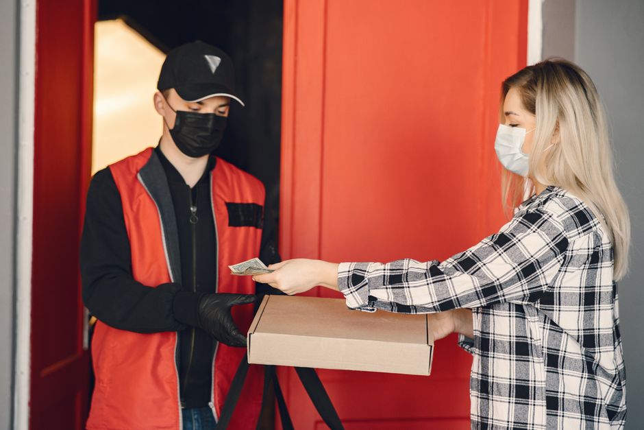 A female customer with blonde hair, wearing a white face mask and a black and white checkered shirt, hands over cash to a courier dressed in a black baseball cap, black face mask, black gloves, and a 