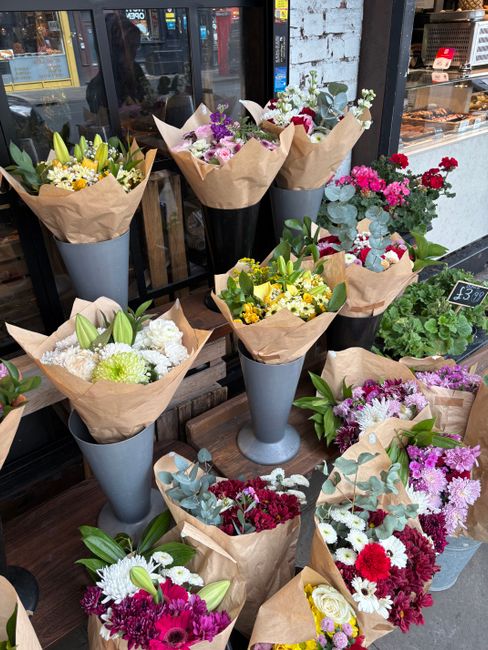 A display of freshly cut floral bouquets arranged in tall, conical grey vases and wrapped in light brown craft paper. The bouquets feature a variety of colorful flowers including white lilies, yellow 