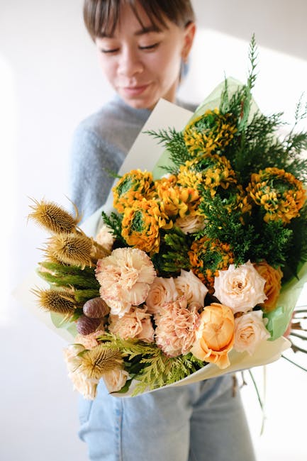 A young woman holding a large, vibrant bouquet of fresh flowers, featuring a mix of peach roses, yellow and orange spray chrysanthemums, green foliage, and textured seed pods. The bouquet is wrapped i