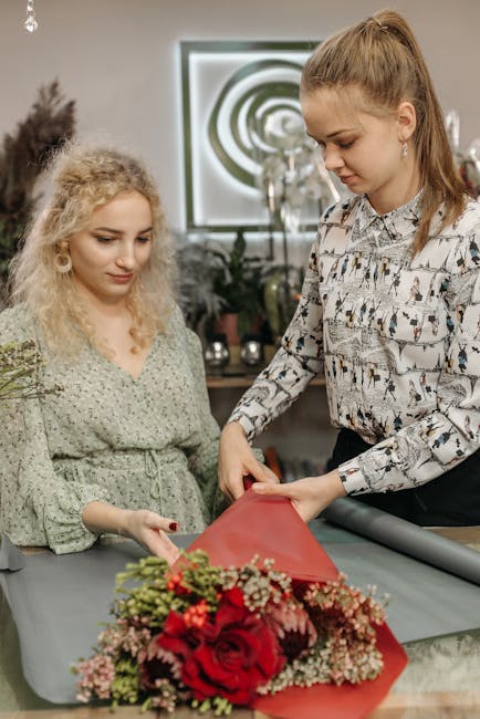 A floral designer at flowersmitcham.co.uk is carefully wrapping a bouquet of vibrant red roses, pink and white baby’s breath, and green foliage in red tissue paper, preparing a fresh floral gift. Th