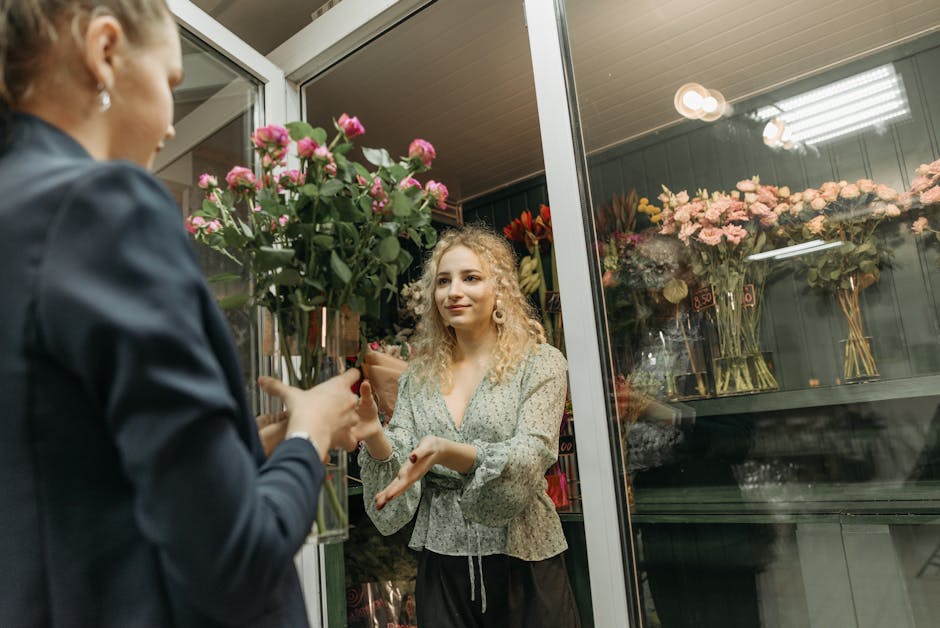A woman with curly blonde hair, wearing a light-colored floral blouse, is engaged in a floral arrangement conversation with a florist inside a flower shop. The florist, dressed in a dark coat, is hand